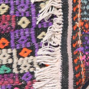 May include: A close-up of a colourful woven rug with fringe. The rug is made of wool and has a geometric pattern in shades of purple, orange, pink, green, and white.