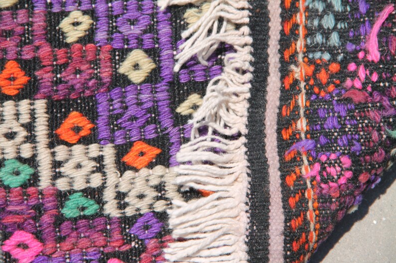 May include: A close-up of a colourful woven rug with fringe. The rug is made of wool and has a geometric pattern in shades of purple, orange, pink, green, and white.