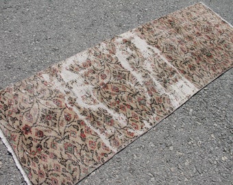 Può includere: Un tappeto da corridore vintage con un motivo floreale sbiadito in tonalità di marrone, crema e rosso. Il tappeto ha un bordo a frangia ed è realizzato in un materiale morbido e tessuto.