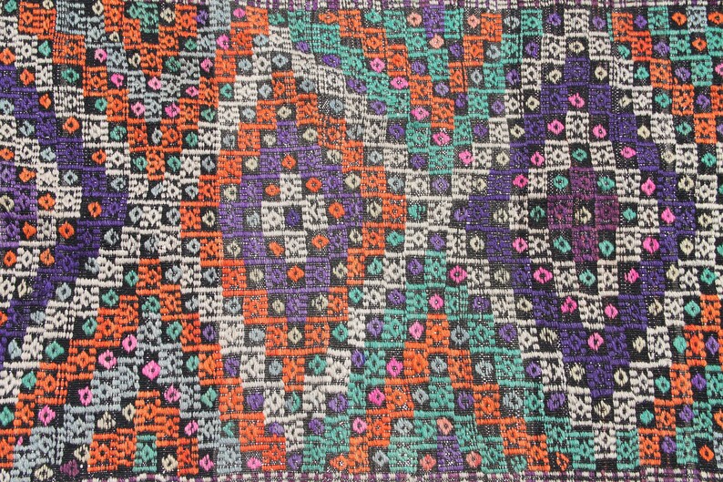 May include: A close-up of a colourful woven rug with a geometric pattern. The rug features shades of orange, blue, green, purple, and white.