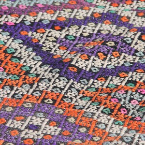 May include: Close-up of a colourful woven fabric with a geometric pattern. The fabric is made of a variety of colours, including orange, purple, blue, and green.