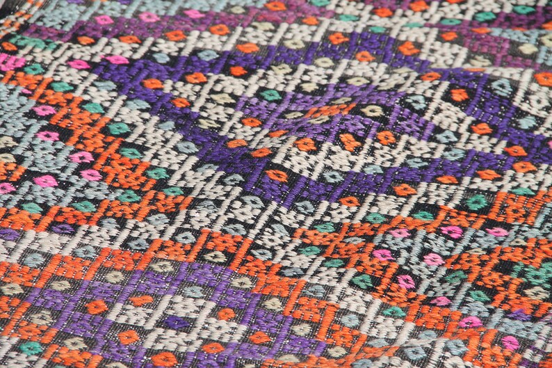 May include: Close-up of a colourful woven fabric with a geometric pattern. The fabric is made of a variety of colours, including orange, purple, blue, and green.