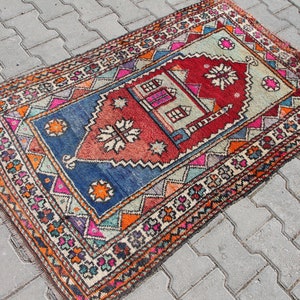 Pu&ograve; includere: Un tappeto turco vintage colorato con un motivo geometrico e un disegno centrale che presenta una moschea e una stella. Il tappeto ha una combinazione di colori blu, rosso, arancione e verde.