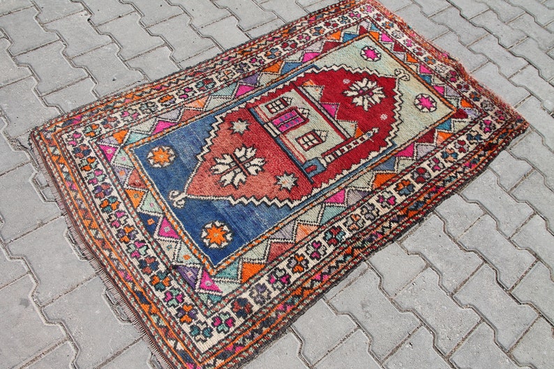 Pu&ograve; includere: Un tappeto turco vintage colorato con un motivo geometrico e un disegno centrale che presenta una moschea e una stella. Il tappeto ha una combinazione di colori blu, rosso, arancione e verde.