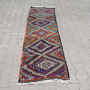 May include: A long, narrow rug with a geometric pattern in shades of orange, purple, green, and white. The rug has a fringe on both ends.