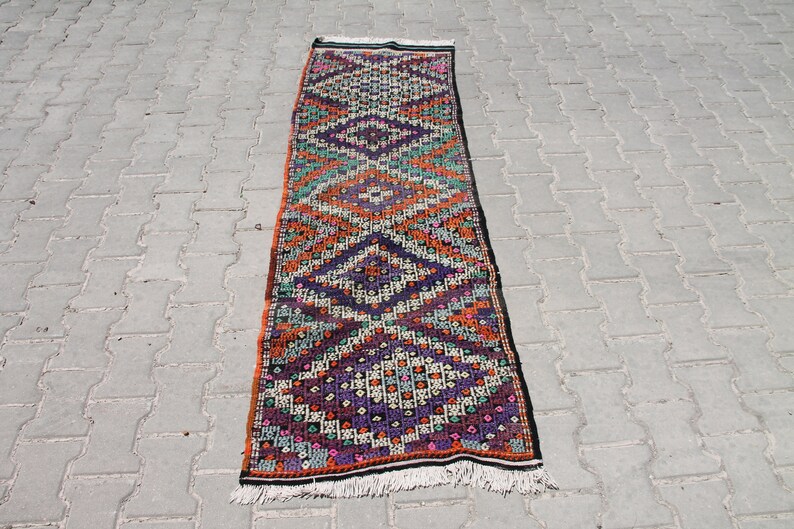 May include: A long, narrow rug with a geometric pattern in shades of orange, purple, green, and white. The rug has a fringe on both ends.