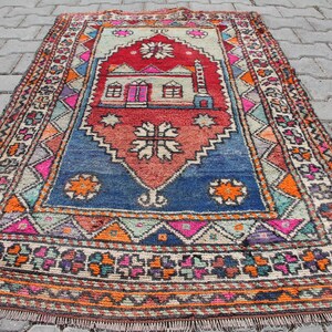 Pu&ograve; includere: Un tappeto turco vintage colorato con un motivo geometrico e un disegno centrale di una casa. Il tappeto &egrave; in tonalit&agrave; di rosso, blu, arancione e verde.