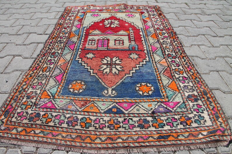 Pu&ograve; includere: Un tappeto turco vintage colorato con un motivo geometrico e un disegno centrale di una casa. Il tappeto &egrave; in tonalit&agrave; di rosso, blu, arancione e verde.
