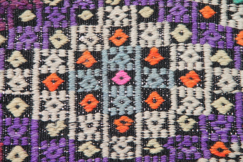 May include: Close-up of a colourful woven fabric with a geometric pattern. The fabric is primarily black with purple, orange, white, and blue accents.