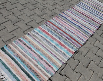 Può includere: Un tappeto lungo e stretto a strisce multicolori con una frangia a un'estremità. Il tappeto è fatto di tessuto tessuto e presenta una varietà di colori, tra cui bianco, blu, rosa, rosso e verde.