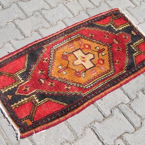 May include: A red, black, and orange patterned rug with a geometric design. The rug is lying on a gray concrete surface.