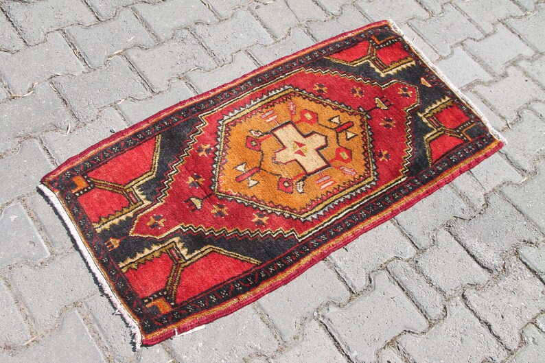 May include: A red, black, and orange patterned rug with a geometric design. The rug is lying on a gray concrete surface.