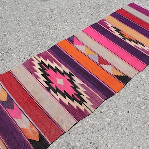 May include: A long, narrow rug with a colourful striped pattern in shades of purple, pink, orange, and red. The rug has a geometric design in the centre, featuring black and white accents.