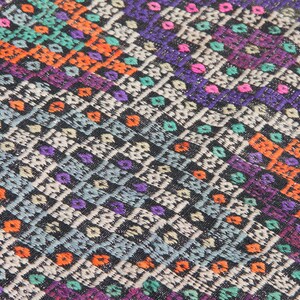 May include: A colourful woven rug with a geometric pattern in shades of orange, purple, blue, green, and white. The rug has a black background and a textured surface.