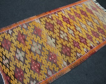 May include: A rectangular, hand-woven rug with a geometric diamond pattern in shades of yellow, brown, and burgundy. The rug has an orange border and white fringe on the ends. The rug is likely made of wool.