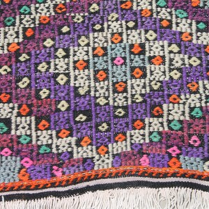 May include: A colourful woven rug with a geometric pattern in shades of purple, orange, green, pink, and white. The rug has a black background and a fringe border.