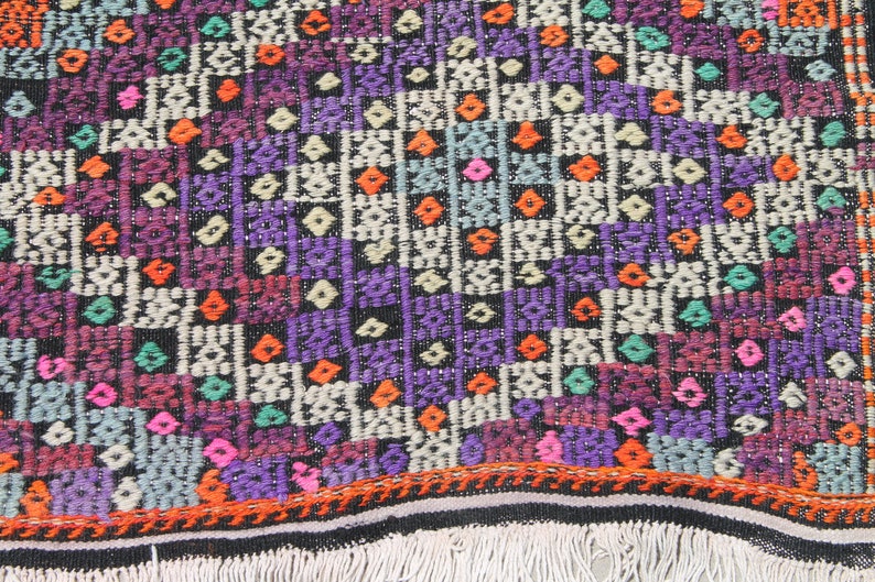 May include: A colourful woven rug with a geometric pattern in shades of purple, orange, green, pink, and white. The rug has a black background and a fringe border.