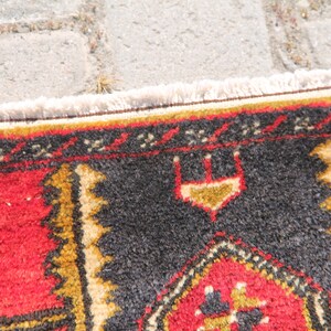 May include: Close-up of a colorful rug with a black, red, and gold geometric pattern. The rug has a white fringe border.