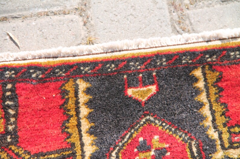 May include: Close-up of a colorful rug with a black, red, and gold geometric pattern. The rug has a white fringe border.