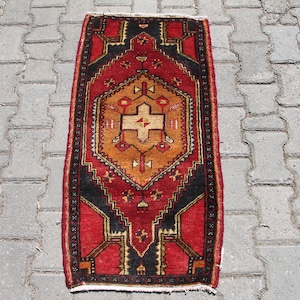 May include: A red, black, and gold patterned rug with a geometric design. The rug is laid out on a gray concrete surface.