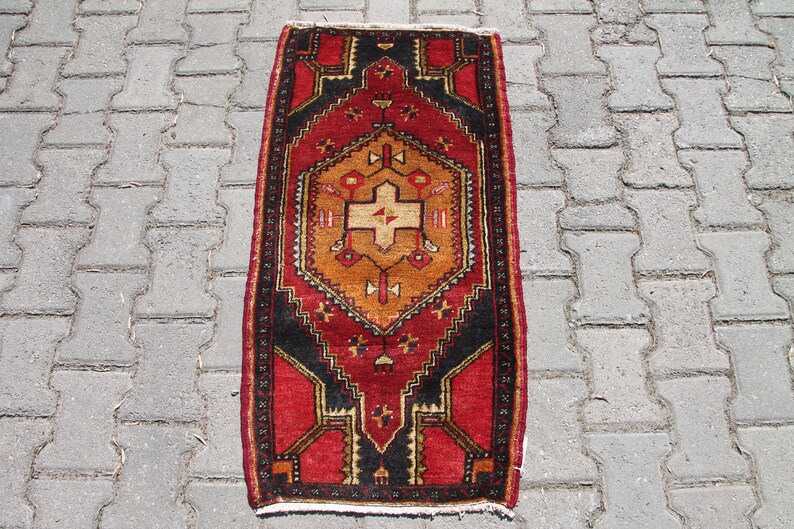 May include: A red, black, and gold patterned rug with a geometric design. The rug is laid out on a gray concrete surface.
