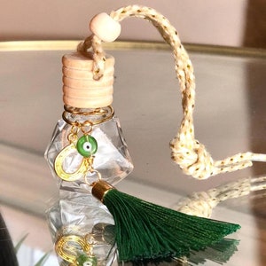 May include: A clear glass car air freshener with a wooden cap and a gold wire accent. It features a green tassel and a charm with a green evil eye and a gold horseshoe. The air freshener hangs from a beige and gold rope.