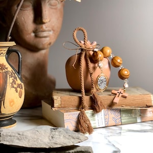 May include: A rosary with large wooden beads and a small wooden cross. The rosary is draped over a stack of vintage books. A small terracotta vase with a rope tie and tassels is in the background, along with a classical bust and a decorative vase.