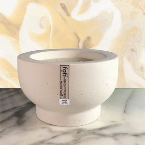Puede incluir: Un portavelas de hormigón blanco con una vela blanca en su interior. El portavelas tiene una etiqueta que dice "föffi - BRIGHT WORKS - vela perfumada - vainilla francesa 12oz/340g"