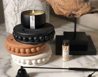 May include: A lit black candle in a metal container sits atop a stack of decorative, beaded candle holders in black, terracotta, and white. A small bottle of matches and a black candle snuffer with a tassel are also present.