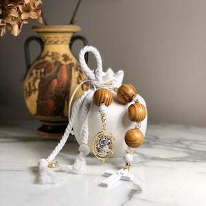 Puede incluir: Un rosario con grandes cuentas de madera, cuentas blancas con aspecto de mármol y un colgante dorado con una imagen religiosa. El rosario tiene una cruz blanca y se exhibe con una bolsa blanca y un jarrón decorativo.
