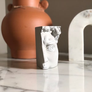 Puede incluir: Una escultura rectangular blanca de un busto masculino clásico, con rasgos faciales detallados. La escultura está colocada sobre una superficie de mármol, con un jarrón de terracota y un objeto blanco en forma de arco en el fondo. Es un objeto decorativo.