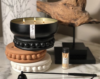 May include: A black three-wick candle with the text "fofi" and "bright works" on a label, sitting atop a stack of decorative candle holders in black, terracotta, and white. A small glass bottle of matches and a black candle snuffer are also present.