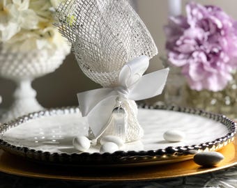May include: White favor bags filled with white almond candies, tied with a white satin ribbon and a silver charm. The bags are on a white plate with a gold rim.