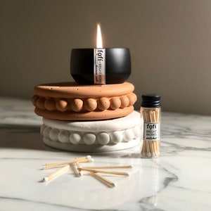May include: A black candle with a lit wick sits atop a stack of decorative, round, terracotta-colored and white stands. A small glass bottle of matches and scattered matches are also present. The candle and matches have the text "fofi - bright works".