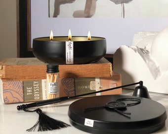 May include: A black, three-wick candle with a gold interior, labeled "fofi". The candle sits on a stack of books next to a match bottle and a candle snuffer. A black lid with a seal and a tassel are also present.