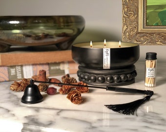 May include: A black three-wick candle in a black ceramic container with a decorative beaded base. The candle is labelled "fofi - bright works" and is surrounded by pine cones and a black candle snuffer with a tassel.