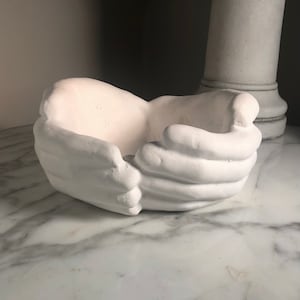 Può includere: Vaso da fiori in ceramica bianca a forma di due mani giunte. Il vaso è appoggiato su una superficie di marmo bianco.