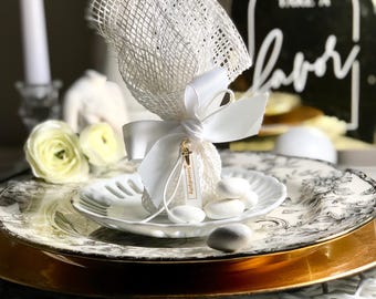 May include: White favor bags filled with white sugar almonds, tied with a white ribbon and a small tag that says "Serendipity". The bags are on a white plate with a gold rim, and a black sign that says "Please take a favor" is in the background.