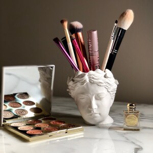 Puede incluir: Un recipiente blanco en forma de busto contiene brochas de maquillaje. Se ve una paleta de sombras de ojos dorada abierta con varios tonos. También hay un pequeño frasco de perfume con tapa dorada.