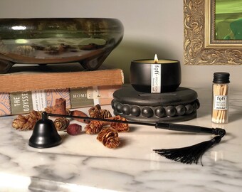 May include: A black candle with a white label that reads "fofi - bright woods" is on a black pedestal. A black candle snuffer with a tassel is on a marble surface with pine cones and other decorative items. A small glass jar with matches that reads "fofi - bright woods matches" is on the right side of the image.