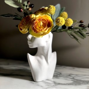 May include: A white ceramic bust vase with a yellow flower arrangement. The vase is shaped like a woman's head and neck with a necklace.