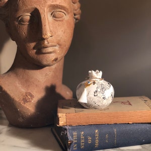May include: White ceramic pomegranate with a gold crown and a white tassel. The pomegranate has a black and white marbled design and a blue evil eye charm. The pomegranate is sitting on top of two books.