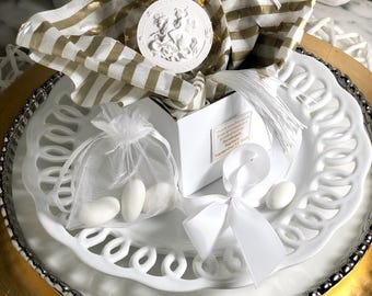 May include: A white and gold themed gift set, featuring a white box with a ribbon, a small mesh bag with white candies, and a decorative medallion. The set is arranged on a white plate with a gold rim, and gold and white striped tissue paper.