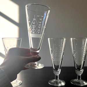 Peut inclure: Ensemble de verres à pied en verre transparent avec des motifs de voiliers gravés. L'ensemble comprend quatre verres, dont un est tenu par une personne. Les verres ont une forme évasée et un pied. L'arrière-plan est de couleur neutre.
