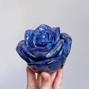 Puede incluir: Una rosa de resina azul con brillo iridiscente. La rosa es un objeto decorativo y está hecha para parecer una flor real.