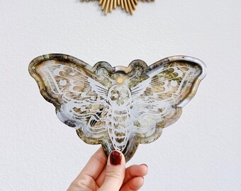 May include: A resin tray shaped like a moth with a white and gold design. The tray is held in a hand with red nail polish.