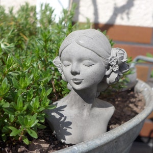 Könnte beinhalten: Eine graue Betonskulptur eines Frauenbuste mit geschlossenen Augen und einer Blumenkrone. Die Skulptur ist teilweise von grünem Laub verdeckt.