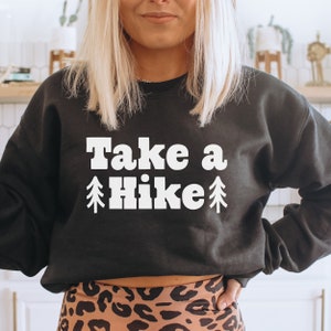 Może przedstawiać: Czarna bluza z białym napisem "Take a Hike" z małym graficznym obrazkiem sosny po obu stronach słowa "hike".