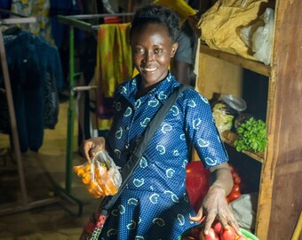 Puede incluir: Una mujer con un vestido estampado en azul y blanco sonríe a la cámara mientras sostiene una bolsa de fruta naranja. Está de pie frente a una mesa con una variedad de frutas y verduras, incluyendo mangos amarillos, aguacates verdes, tomates rojos y fruta naranja.