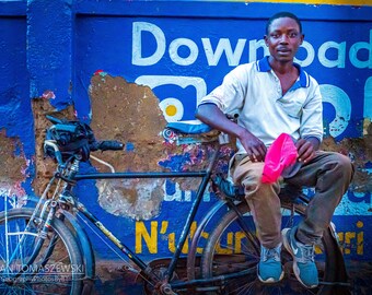 Puede incluir: Un hombre con una camisa polo blanca y pantalones caqui se sienta en una bicicleta con un fondo azul y amarillo. El fondo tiene el texto "Downloading" en él.